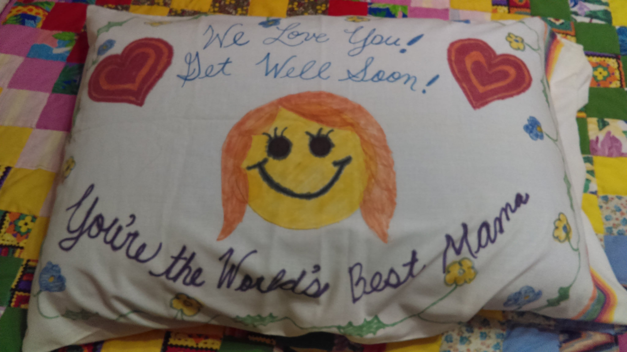 The front of the handmade pillow, with a smiling face, hearts, and the words “We Love You! Get Well Soon! You’re the World’s Best Mama.”